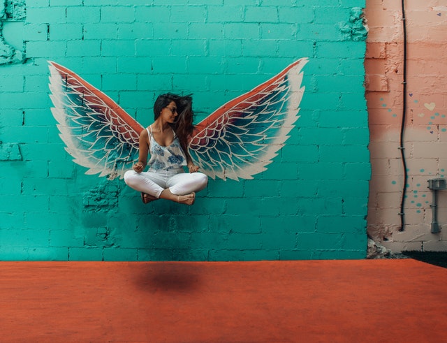 Anjelské krídla namaľované na tehlovej stene, pred ktorými sedí žena v tureckom sede